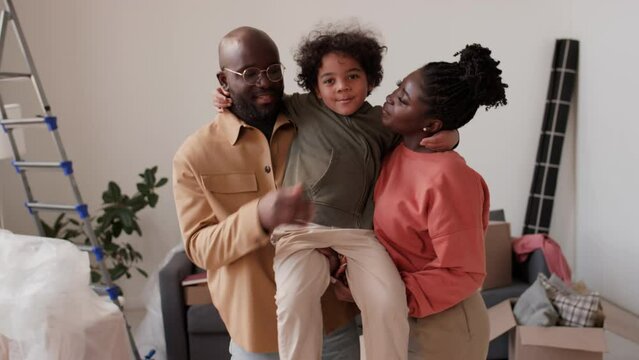 Portrait of joyous African American parents holding little son on arms and posing together for camera with smile in new house with unpacked stuff after moving