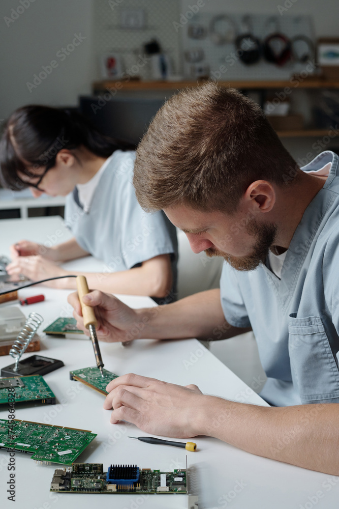 Poster young man with electric handtool bending over desk and motherboard while repairing detail of compute