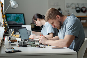 Professional young technician bending over desk while repairing or checking detail of computer motherboard against female colleague