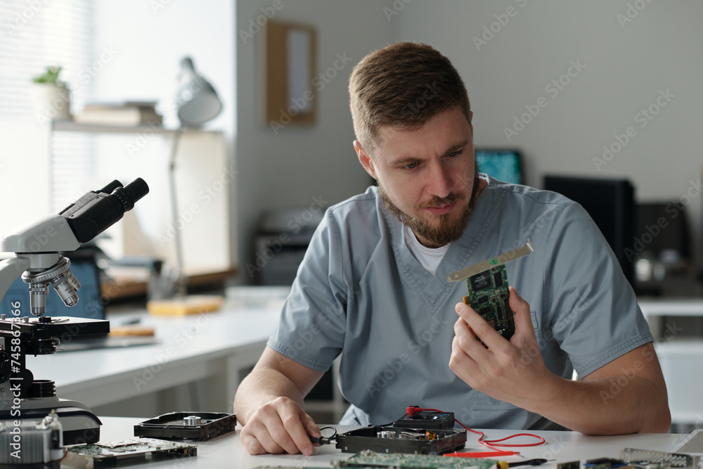 Poster Young serious man in grey uniform looking at part of computer processor while sitting by workplace and identifying technical problem