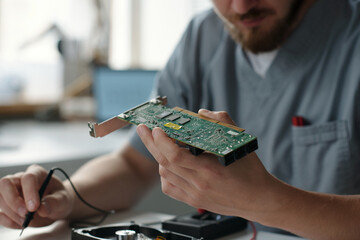 Hand of young repairman in uniform holding motherboard while sitting by workplace in repair service...