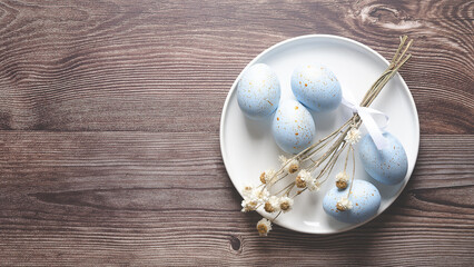 Blue painted Easter eggs with golden spots and some dry flowers  on a plate with flowers on wooden background and copy space.