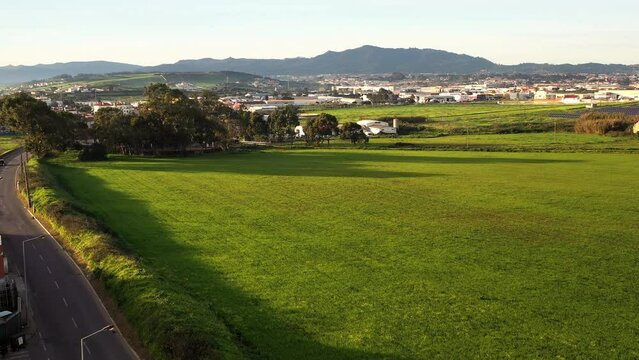 Aerial drone captured tilt-down shot of green meadow landscape, with vehicles driving on asphalt road, Sintra town in the background, all under clear sky on sunny summer day.