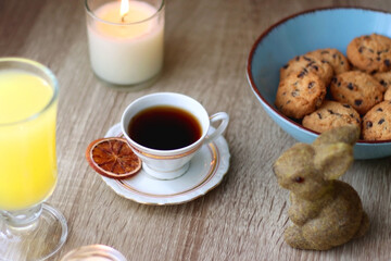 Easter eggs in the basket, bowl of cookies, chocolate pralines, Easter bunny figurine, cups of tea, glasses of juice, flowers and lit candles on the table. Selective focus.