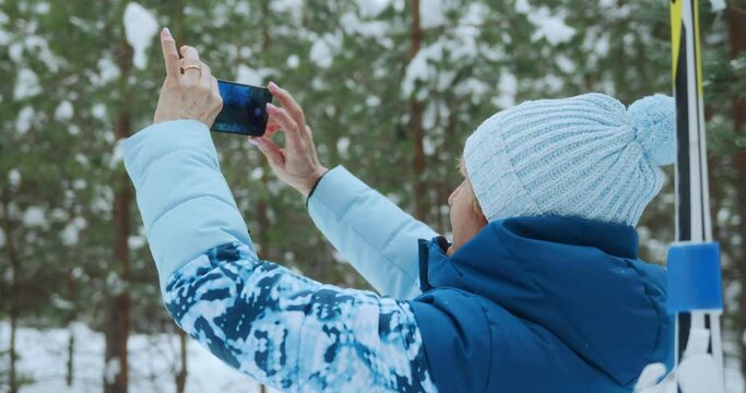 A 65-year-old adult female skier in a blue ski suit walks in a winter forest and takes photos of snow trees using her phone or smartphone. High quality 4k footage
