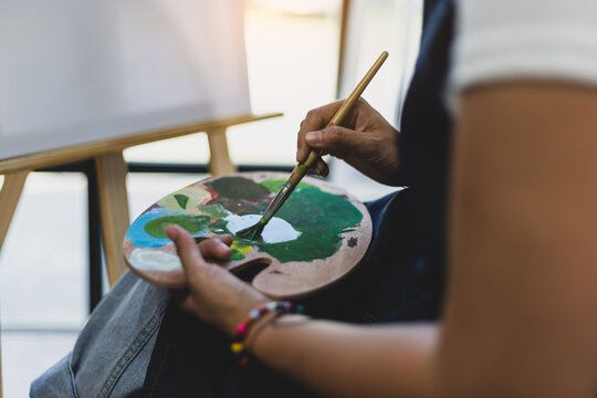 Artist or female painter holding a palette and paintbrush paints and paints her artwork. Young Asian woman drawing in her free time at home.