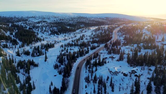 Aerial drone footage of snow covered mountains in Sweden, with small village and winding road towards setting sun, sunset. Many pine trees and forest covered in white snow on hills.