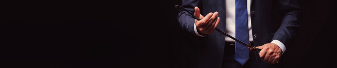 man dominant boss in a suit holds a leather flogger whip in hands for BDSM sex with domination and submission. Wide horizontal banner header with a copy space on black background - obrazy, fototapety, plakaty
