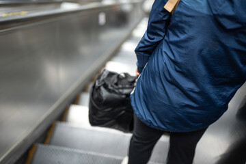 Close-up at behind of a person holding bag is standing on the escalator during moving down to the...