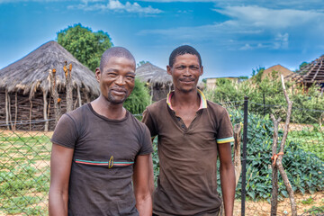 two village young and happy african men standing in front of a thatched hut.
