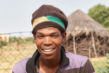 village young african man with a beanie standing in front of a thatched hut.