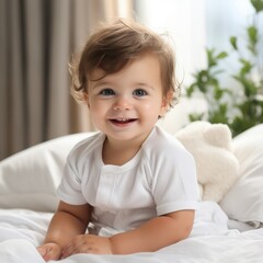 Happy baby boy sitting on a bed with white blankets. Beautiful smiling infant in a white shirt sitting on a bed in a light bedroom. Cute cheerful baby boy looking at the camera.