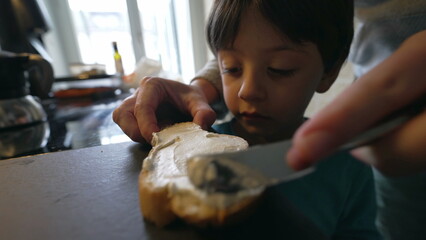 Child looking at parent's hand spread cream cheese on bread's surface, close-up small boy face...