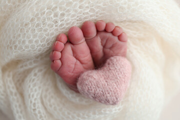 The tiny foot of a newborn baby. Soft feet of a new born in a white wool blanket. Close up of toes, heels and feet of a newborn. Knitted pink heart in the legs of a baby. Macro photography