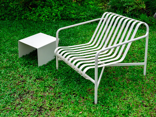 Empty modern minimal design of white empty curved armchair seat and small square side table, iron materials, decoration on green grass background at the summer outdoor backyard garden with nobody.