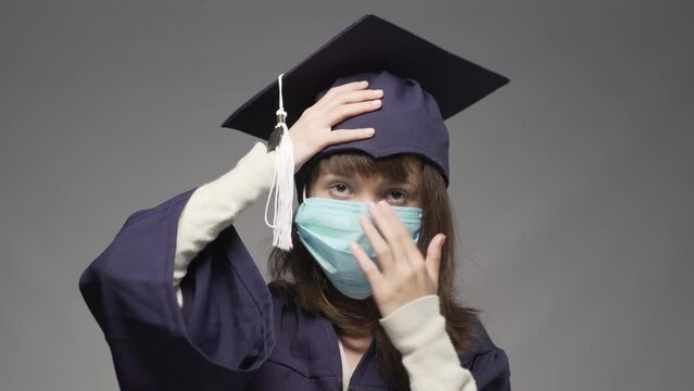Graduation girl wearing cap and protective face mask. Female student posing against gray background.
