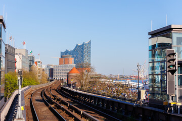 Obraz premium Hamburg Red Unmanned U-bahn Subway train rail Baumwall station on Elevated Track with Hamburger Port Harbor Elbphilharmonie building background blue sky. Hanseatic city commute transport scenic view