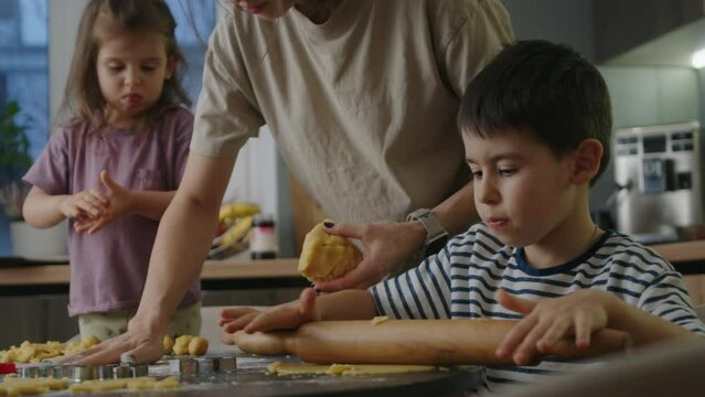 Little boy rolling dough while mother teaching little girl daughter pressing cookie's dough using hands. Child development, learning, mom teaching kids cooking