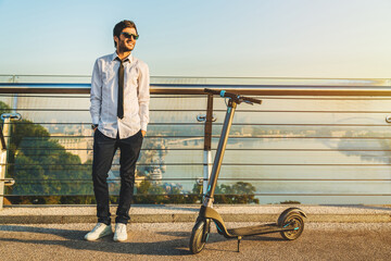 Stylish male in sunglasses posing near electric scooter on modern bridge with city view on...