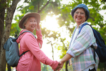 Two elderly women travel in nature, smiling happily Wear hats and backpacks and hold hands. Elderly...