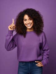 Happy smiling woman with curly hair wearing purple sweater pointing at something with finger isolated on purple background with empty space for text or inscriptions 