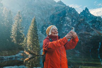 Fototapeta na wymiar Rear shot of traveler 60s woman influencer in sportswear with backpack standing on the rocky shore and making selfie on smart phone. Taking photo of green hills and mountains on Morskie Oko lake