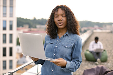 Young beautiful African American female looking at camera while holding laptop computer, standing...