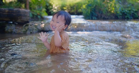Exuberant toddler splashing in a shallow stream, laughter sparkling as sunlight filters through green foliage in a blissful outdoor scene