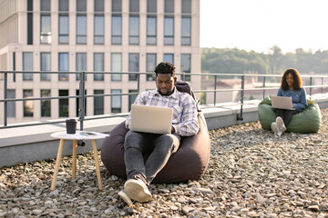 Successful business man dressed in checkered shirt and pants sitting at chair bag on roof top outdoors and typing on modern laptop, African American colleague works sitting in bag chair in background. - Powered by Adobe