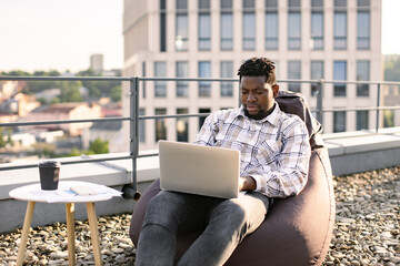 Business man in casual checkered shirt working on laptop. Professional businessman using digital device having video chat conference, sitting on beanbag on rooftop of modern building. - Powered by Adobe