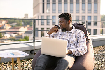 Young man in casual wear carrying out professional activities in open air. Portrait of African male drinking coffee while placing laptop on knees sitting on bean bag chair on roof terrace. - obrazy, fototapety, plakaty