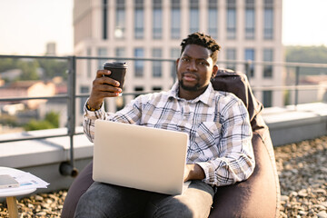 Adult businessman in casual wear carrying out professional activities in open air. Portrait of African male drinking coffee while placing laptop on knees sitting on bean bag chair on roof terrace.