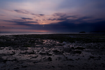 Fototapeta premium Anochecer con sol detras de las nubes y luces rojizas i violetas con algas verdes en Ujung Genteng, Sukabumi, Java, Indonesia