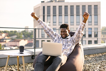 Portrait of African male drinking coffee while raising hand up, celebrating good news sitting on...