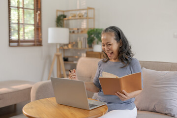 Excited senior Asian woman holding notebook celebrating success with laptop at home Online success and happiness concept.