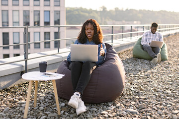 Focused African female wearing denim shirt and headset holding online conference over portable computer on rooftop, male colleague working on blurred background.