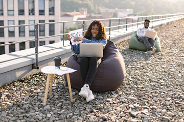 Calm African American businesswoman in denim shirt and headset discussing financial data plan on panoramic terrace with male colleague working on blurred background.