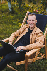 Cute man in a shirt sitting in a chair outdoors in the garden and working on a laptop, remote work, female freelancer