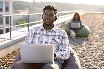 Smiling African American man in casual clothes looking at camera using portable computer and...