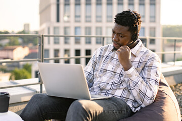 Young African man in casual clothes using portable computer and headset in cozy beanbag on roof terrace. Well-organised employee enjoying open air while staying available during midday. - obrazy, fototapety, plakaty