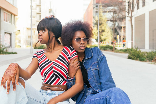 Two sisters share a moment on a city sidewalk, their casual styles and relaxed postures capturing the essence of urban life