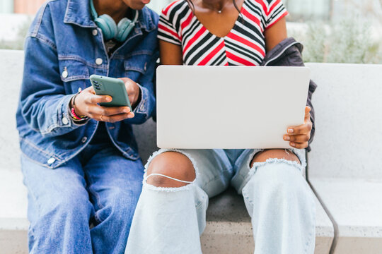 Two anonymous young women, possibly sisters, are engrossed in their digital devices, sharing content and connectivity in a casual urban setting