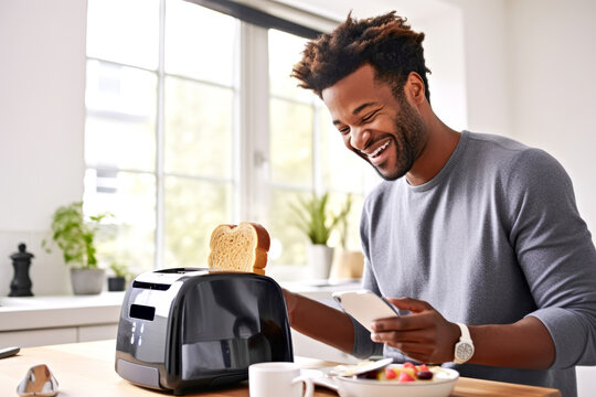 Cheerful smiling african american man uses smartphone app to control a modern black smart toaster in bright sunny kitchen. Concept of convenience wireless modern technology for cooking meal breakfast