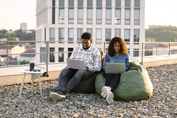 Beautiful young African American couple using laptops while sitting in beanbags at rooftop of...