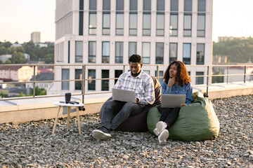 Beautiful young African American couple using laptops while sitting in beanbags at rooftop of...