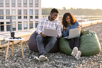 Smiling African couple sitting at beanbags at rooftop of office with laptops and discussing project...