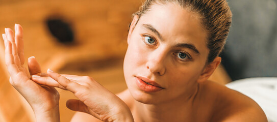 A portrait of beautiful young woman lies on spa bed in front of wooden sauna cabinet. Attractive caucasian women in white towel looking at camera with relaxing environment. Close up. Tranquility.