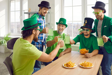 Group of a happy smiling male friends celebrating Saint Patrick's Day at home. Joyful men in green hat and glasses having fun at the party drinking beer with french fries and chips.