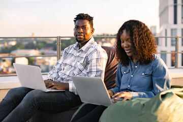 African colleagues working with laptops while sitting at beanbags in modern rooftop terrace of office. Portrait of happy businessman posing to camera near her female colleague.