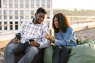 African male and female colleagues sitting on chair bags on rooftop and using smartphones for...